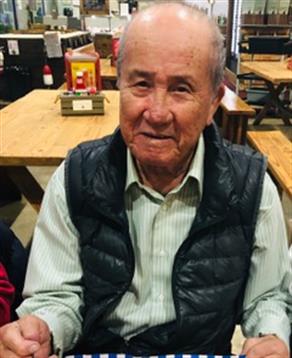 An elderly man smiles while holding a plate of food in a bustling restaurant setting.