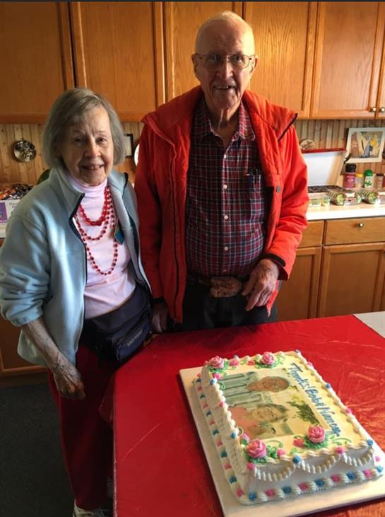 Elderly couple enjoys a special moment together, standing beside a decorated cake in a warm kitchen.