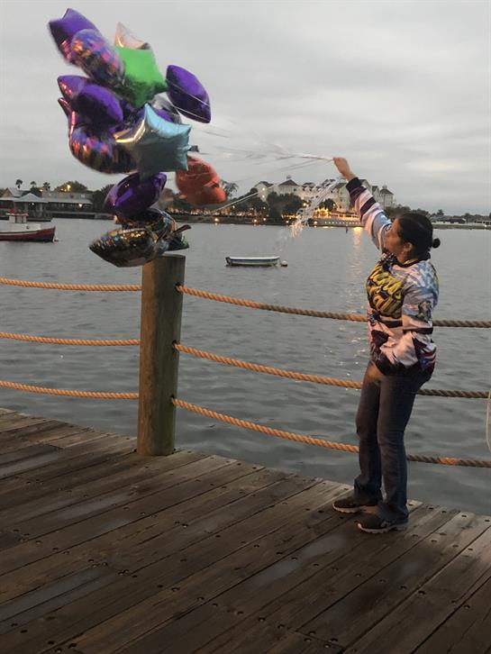 A young girl stretches out her arm to catch shining balloons drifting above a dock on a cloudy day.