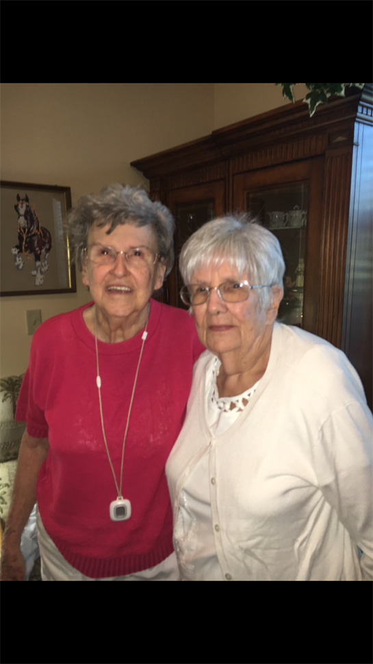Two older women stand side by side, smiling warmly in a comfortable living space filled with decor.