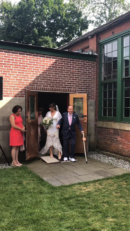A bride and groom joyfully leave their wedding venue, greeted by a guest.