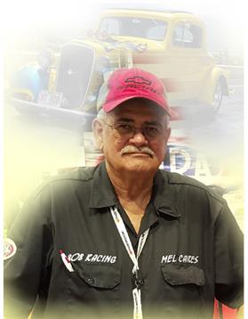 A man wearing a red cap and dark shirt stands proudly at a racing event among vintage cars.