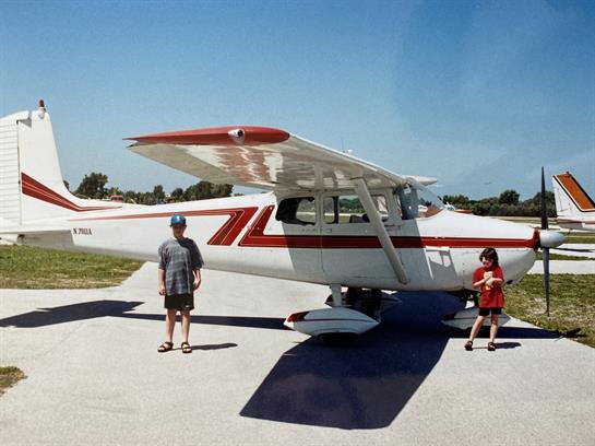 Two children stand next to a white and red aircraft on a bright, clear day at an airfield.