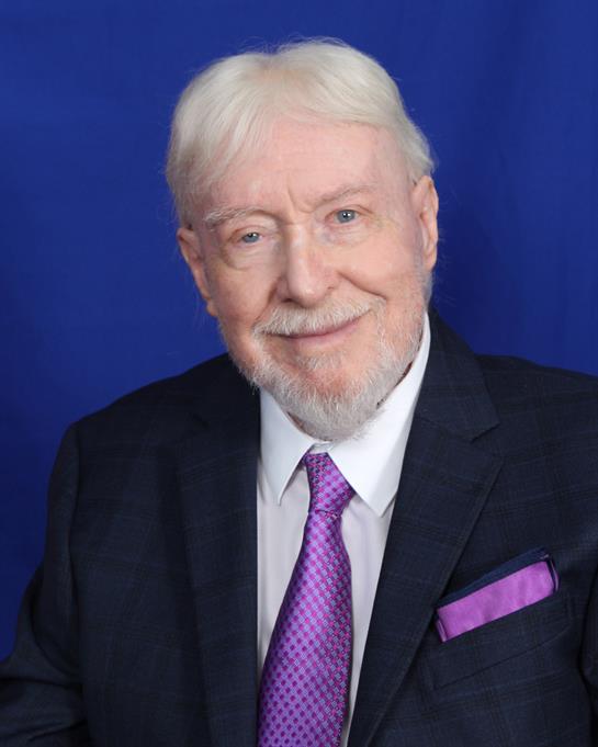 A smiling elderly man in a suit and tie stands against a bright blue background.