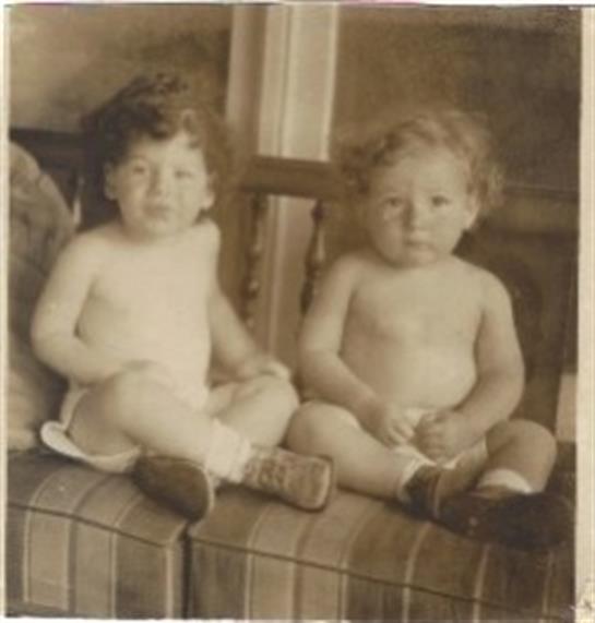 Two adorable toddlers sit side by side on a striped couch, smiling and playing in a warm atmosphere.
