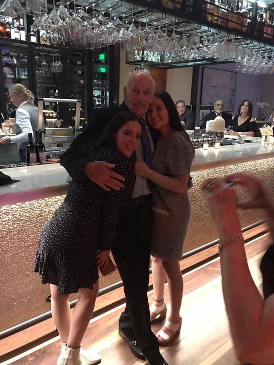 Two women are smiling and posing closely with a man at a bar during a vibrant evening gathering.