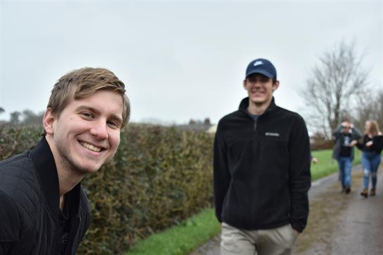 Two young men walk together along a rural path surrounded by hedges under a cloudy sky.