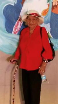 An elderly woman wearing a vibrant red blouse stands next to a colorful mural with a cane.