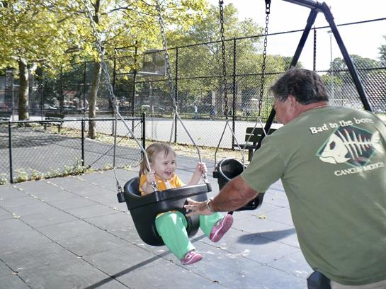 A young child swings joyfully while a caregiver gently pushes them in a busy park.
