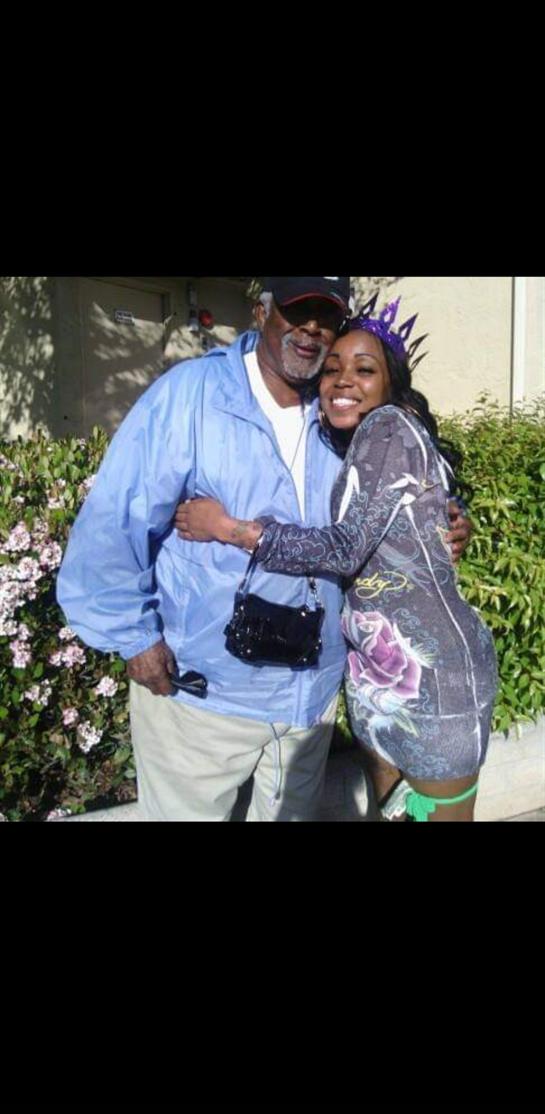 Two people warmly embrace each other in a sunny garden, surrounded by colorful flowers and greenery.