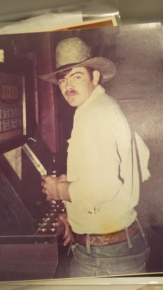 A mustached man in casual western wear plays a slot machine in a rustic bar.