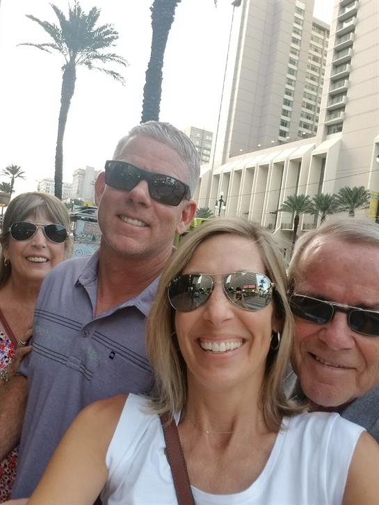 Four friends smile outside, enjoying a sunny day in an urban environment surrounded by palm trees.