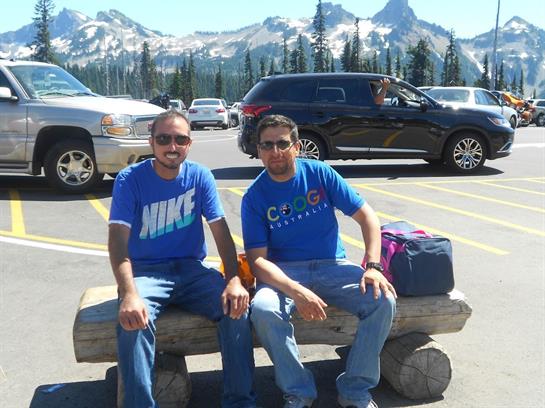 Two friends sit on a log in a parking area surrounded by mountains on a clear day.