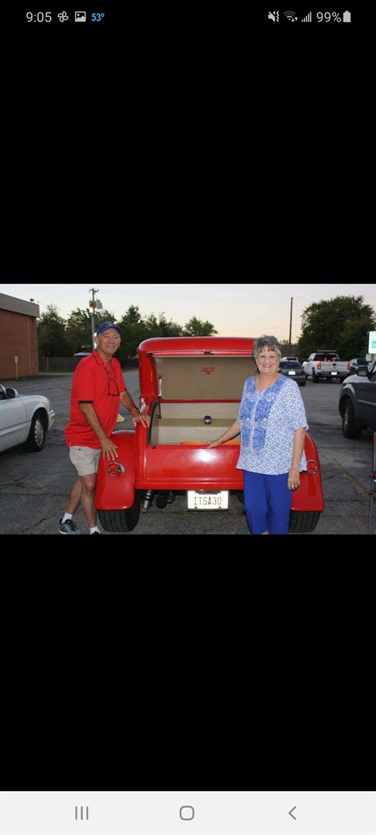 A cheerful couple poses beside a classic red automobile in a parking lot during dusk.