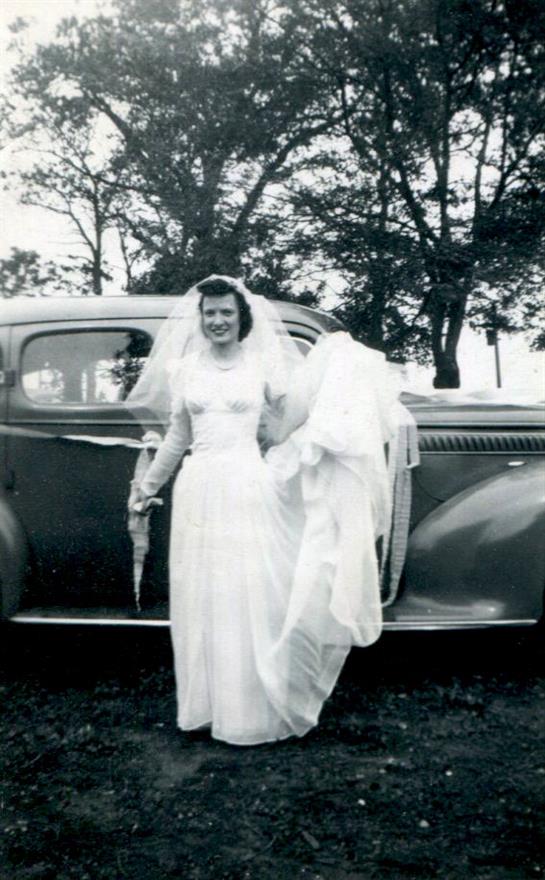A bride in a classic gown and veil holds an umbrella by a vintage car in a sunny rural area.