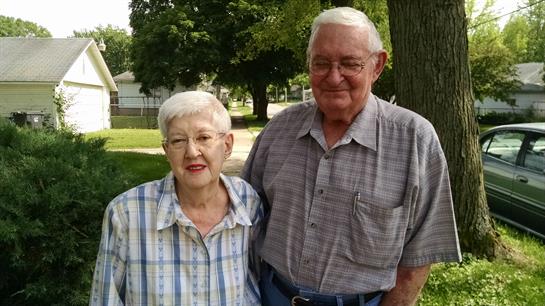 An elderly couple stands side by side, smiling warmly in a green park surrounded by trees.