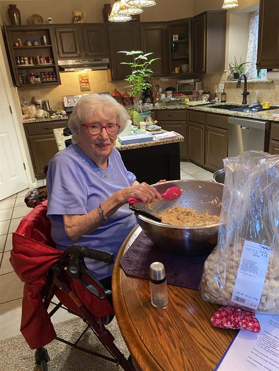 An elderly woman is mixing cookie dough in a large bowl while seated at a kitchen table.