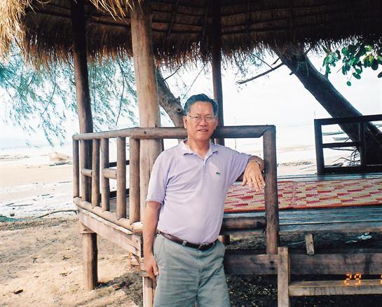 A man stands relaxed by a coastal gazebo surrounded by trees and sand, enjoying the scenic view.