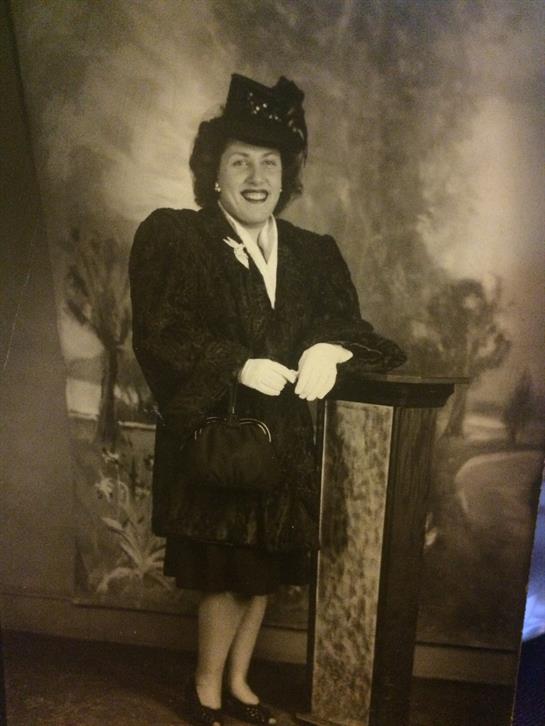 A stylish woman stands with a smile, wearing a vintage outfit and gloves, in an elegant environment.