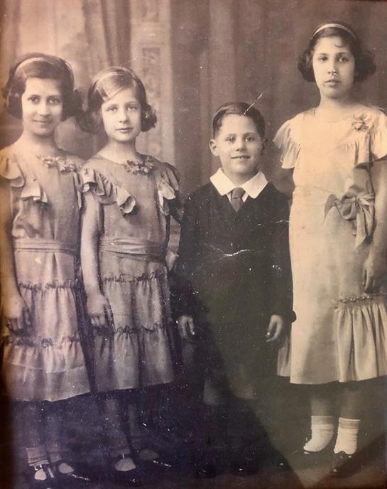 Four children stand side by side in old-fashioned attire, smiling for the camera in a studio.