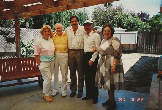 Group of five friends smiles and poses together under a wooden structure in a sunny garden.