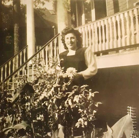 A woman stands in a garden full of flowers, smiling at the camera in natural light.
