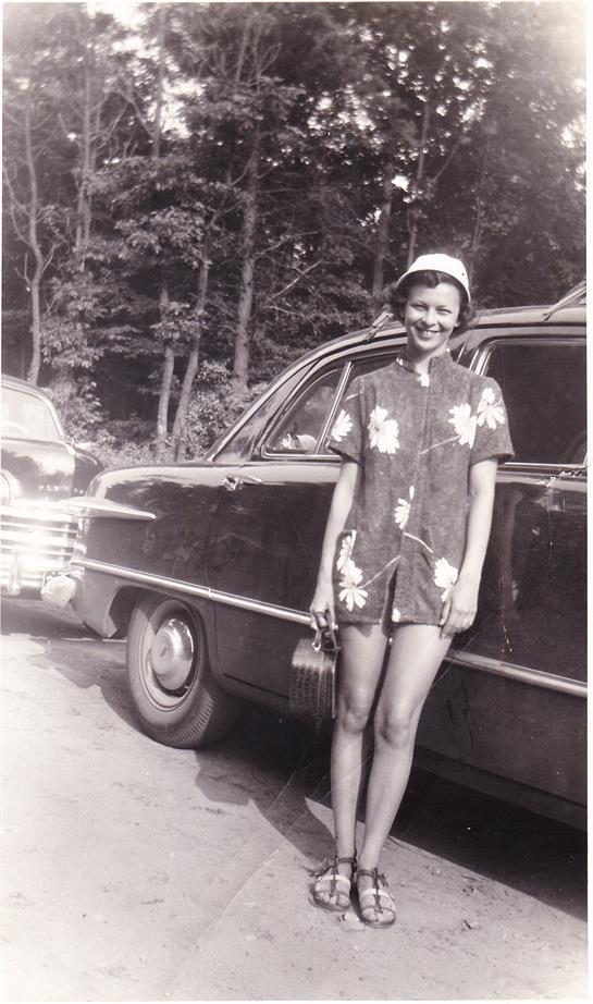 A young woman smiles in a floral outfit beside a vintage car in a scenic forest setting.