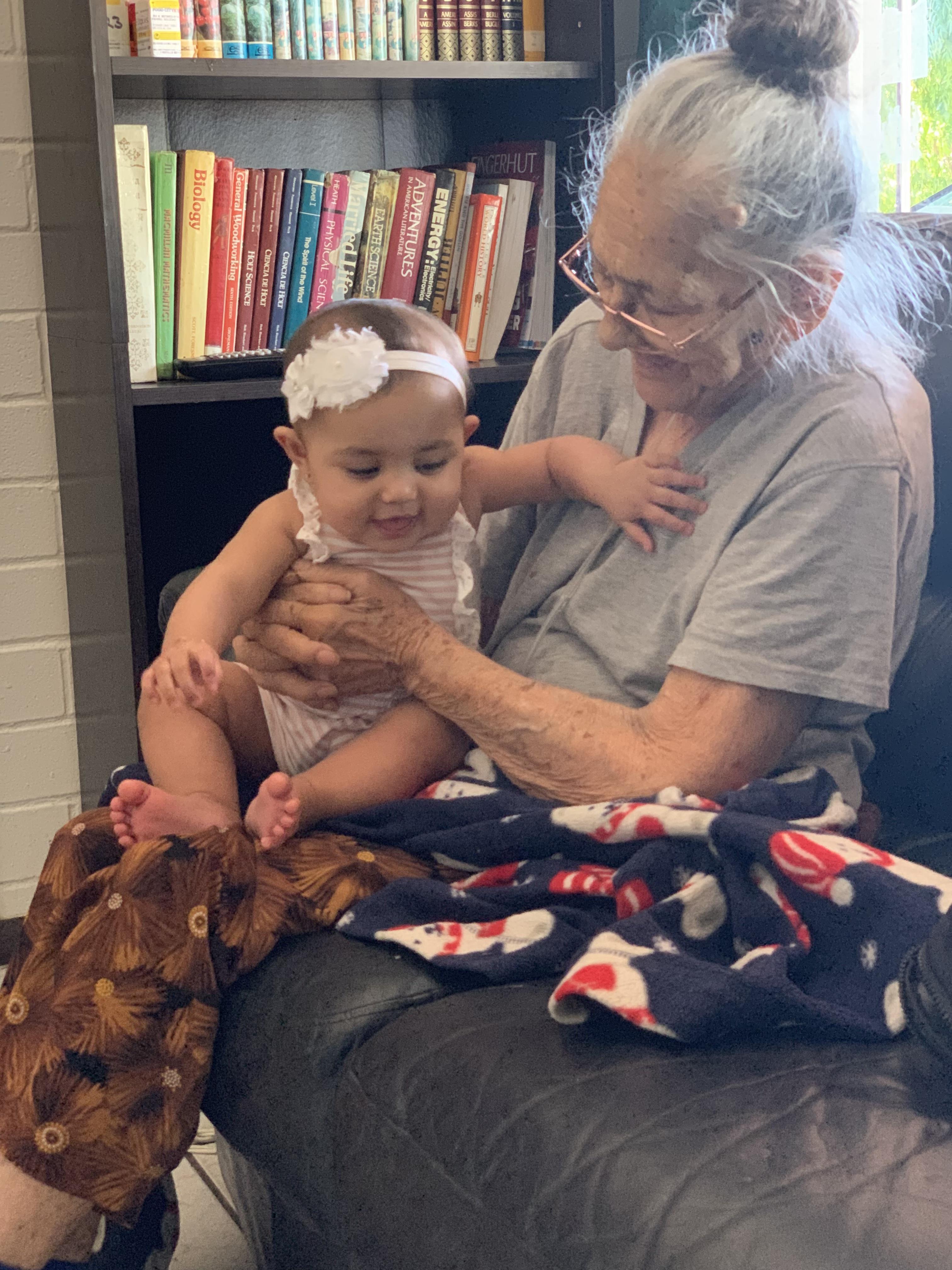 An elderly woman lovingly interacts with a baby on her lap, surrounded by books and warmth.