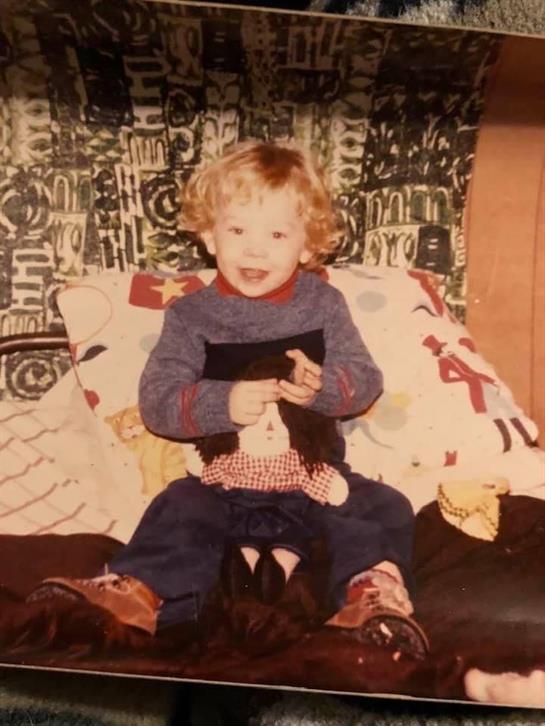 A young child is smiling while holding a doll, sitting on a couch with vibrant fabric decorations.