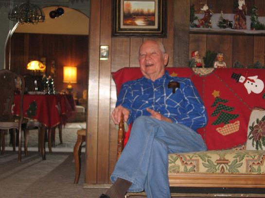 An older man smiles warmly in a cozy, festive holiday living space.