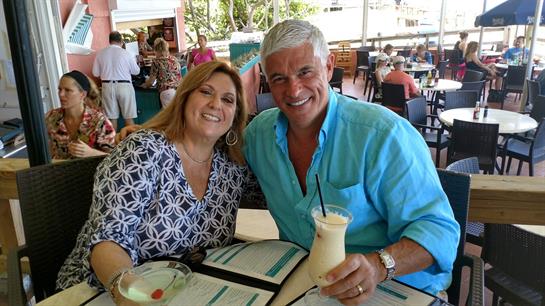 A couple smiles while enjoying tropical drinks at a lively restaurant with friends around.