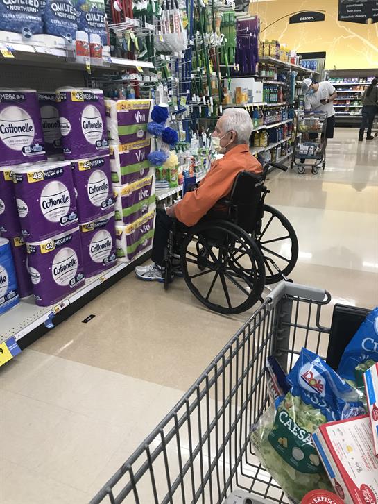 An elderly person in a wheelchair examines products on a store shelf while shopping.
