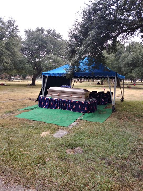 A gathering is taking place near a casket covered by a blue tent in a peaceful cemetery setting.