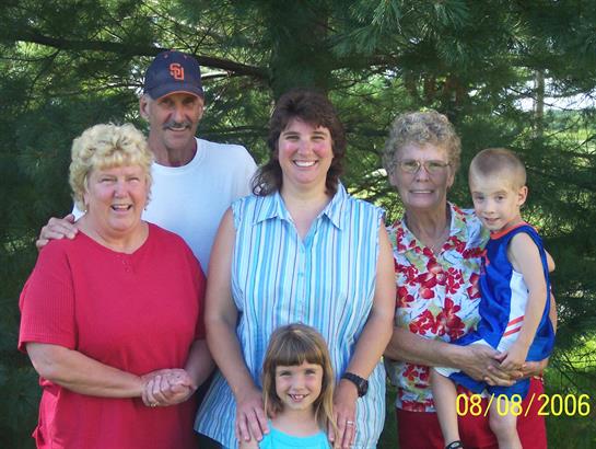 Five family members stand together smiling under green trees in a warm outdoor location.
