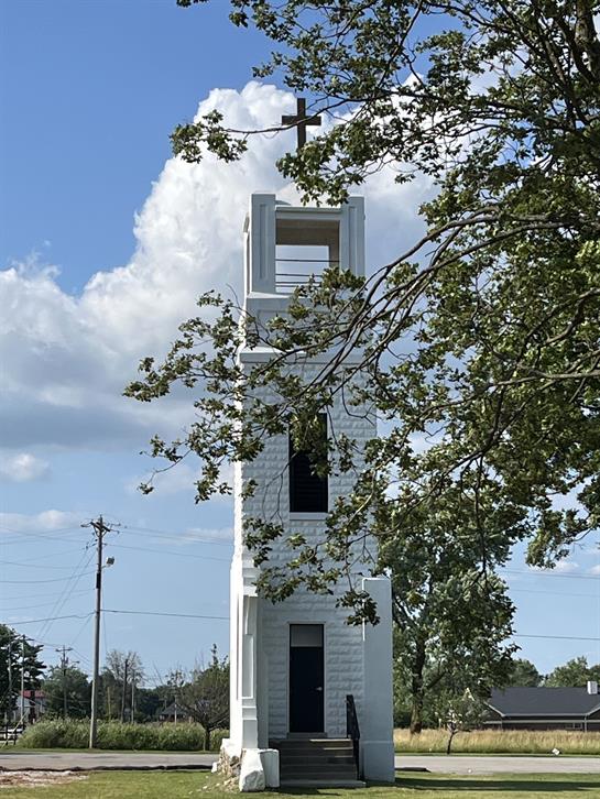 A tall church tower with a cross stands among green trees under a clear blue sky.