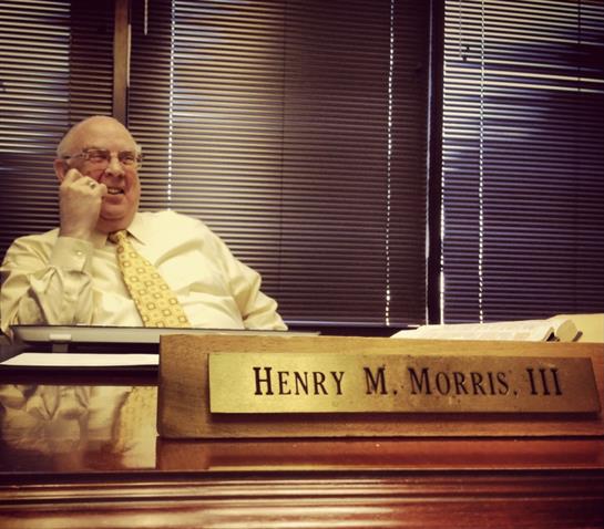 A man in a suit smiles while talking on the phone in an office with books and blinds.