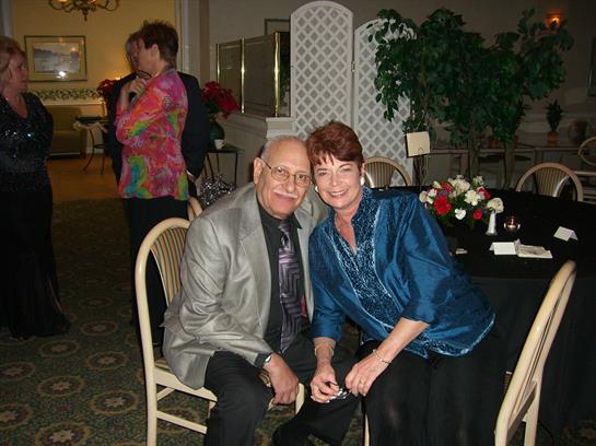 A couple smiles happily while seated at a table during a lively gathering in an elegant setting.