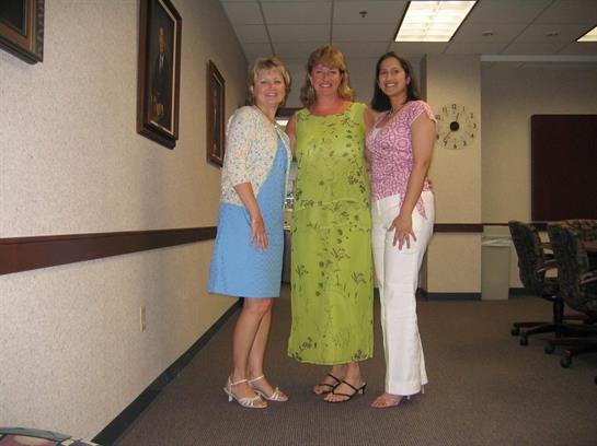 Three women stand together in an office hallway, smiling and dressed in spring attire.