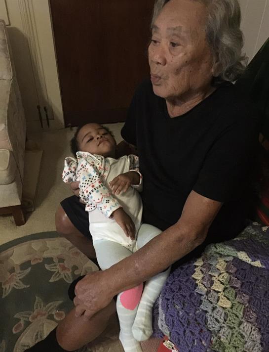 An elderly woman sits on a chair holding a peacefully sleeping baby, creating a warm moment.