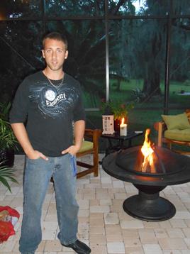 A young man poses casually next to a glowing fire pit in an inviting outdoor space.