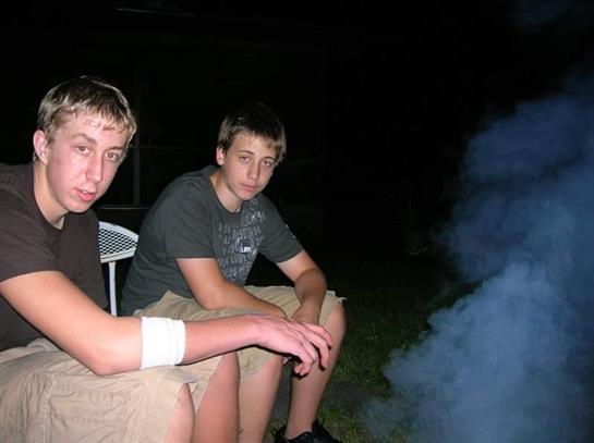 Two teenagers sit together in a backyard, surrounded by smoke from a fire, enjoying their evening.