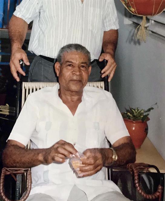 An elderly man sits in a chair with a small object, while someone stands behind him.