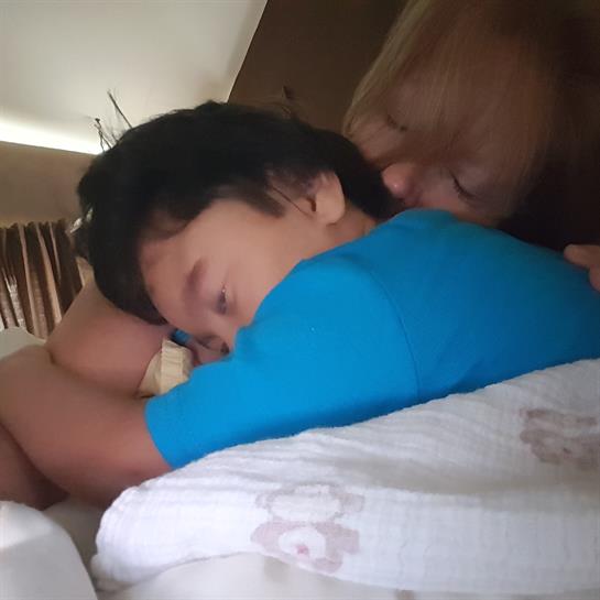 A child is comfortably resting on a bed while a parent gently hugs and kisses them.