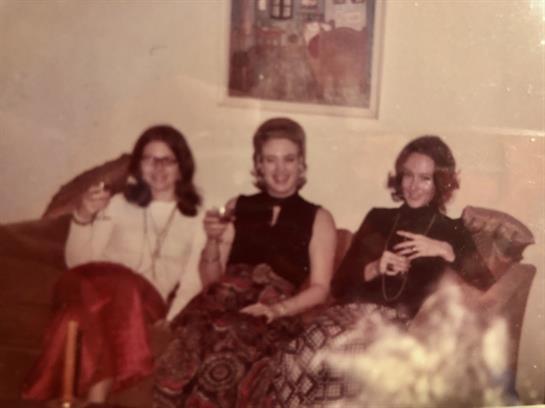 Three women on a couch are smiling and raising their glasses, enjoying time together.