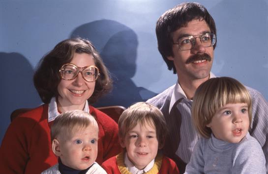 A family sits together, smiling warmly, dressed in vibrant colors typical of the 1980s fashion.