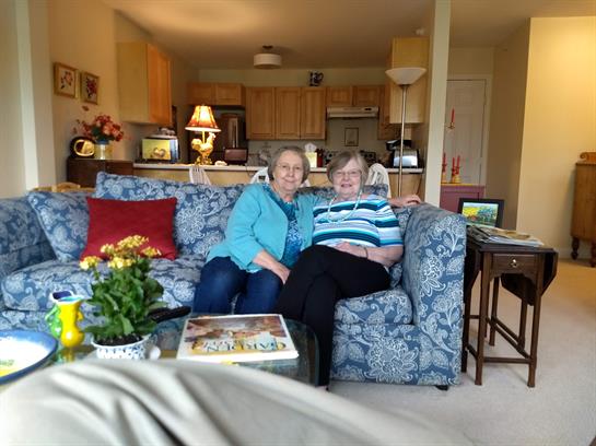 Two women sit closely on a couch, smiling and enjoying each other's company in a sunny living room.