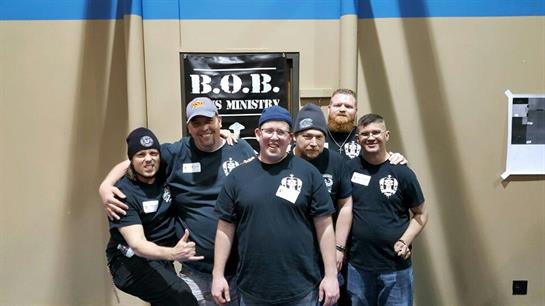 Six men stand together smiling, engaged in camaraderie at a community gathering indoors.