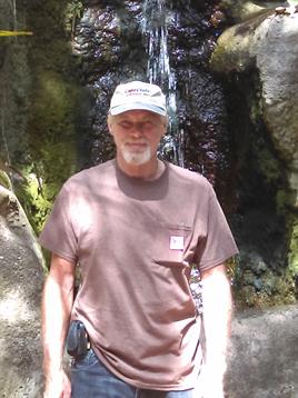 A man wearing a cap stands confidently in front of a scenic waterfall, surrounded by greenery.