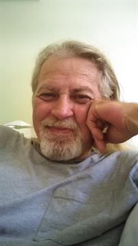 An older man with a gray beard smiles while resting indoors, enjoying a relaxed moment.