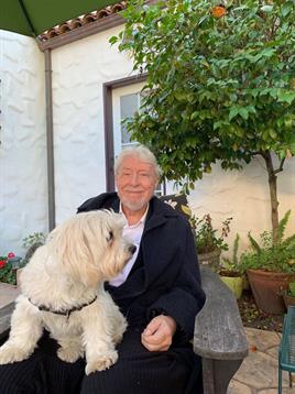 A man sits on a bench with his dog surrounded by plants in a serene outdoor setting.